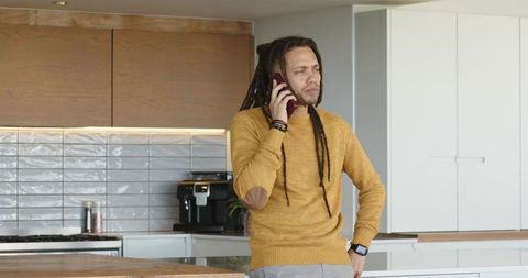 Mid adult man leaning on wooden island in modern kitchen talking on phone in mustard sweater