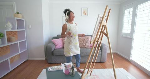 African american woman painting at easel in bright living room wearing paint-stained apron