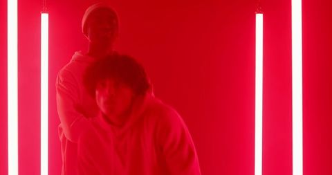 Two African American male colleagues are captured in a striking pose within a creatively lit studio area. Dramatic red LED lighting creates a bold and intense atmosphere, highlighting their urban fashion style with cozy hoodies. This composition conveys a sense of modernity and dynamic energy, making it ideal for fashion campaigns, creative projects, and advertisements focusing on contemporary style and energy.