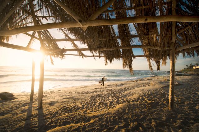 Captures a beautiful sunset scene at a beach with a surfer walking towards the ocean. The foreground shows a rustic wooden pavilion covered with palm fronds, adding to the tropical ambiance. Perfect for promotional materials for surfing, vacation spots, tropical getaways, and relaxation retreats.
