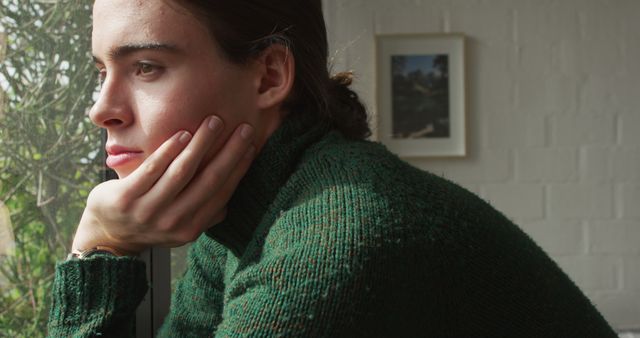 Young woman in green sweater sits by the window, gazing outside with a thoughtful expression. Perfect for themes related to introspection, peaceful solitude, quiet moments, relaxation, and inner thoughts. Can be used in mental health content, lifestyle blogs, and articles on mindfulness or personal reflection.