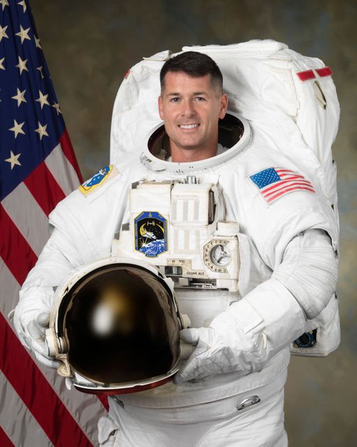 Astronaut posing in a space suit while holding a helmet with the American flag and NASA Johnson Space Center background. Excellent for use in articles or materials related to space exploration, NASA missions, and astronaut profiles. Can be used in educational content about space, promotions for science and technology events, or patriotic displays related to space achievements.