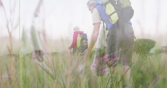 Caucasian couple, both fitness instructors, are hiking through a meadow with backpacks, enjoying the outdoors and nature together. Suitable for images showcasing outdoor adventures, active lifestyle, fitness, healthy living, nature exploration, outdoor activities advertising.