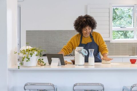 Home baker kneading dough on sunlit kitchen island with tablet, flour jar, milk carafe