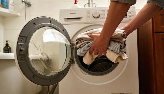 Hands loading towels and striped shirt into front-load washer in bathroom laundry nook