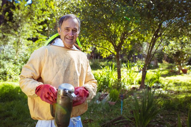 Senior man wearing beekeeper uniform smiling in lush garden. Ideal for use in articles about beekeeping, sustainable living, hobbies for seniors, and honey production. Can be used in promotional materials for beekeeping equipment or environmental conservation campaigns.