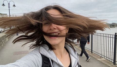 Young woman laughing on riverside selfie with wind-blown hair, hoodie and backpack