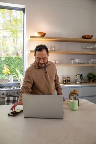 Mature Asian man working from home at kitchen island with laptop and smartphone