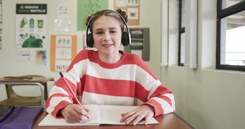 Young Girl in Classroom Writing with Headphones and Notebook