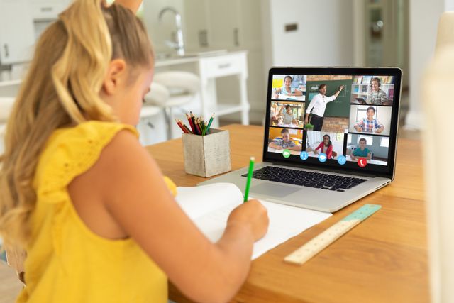 Caucasian girl with blond hair writing in book while learning from online class over video call. Laptop, home, screen, unaltered, childhood, wireless technology, education, student and e-learning.