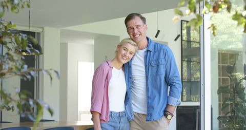 Young couple leaning and smiling at camera in sunlit contemporary home by glass doors