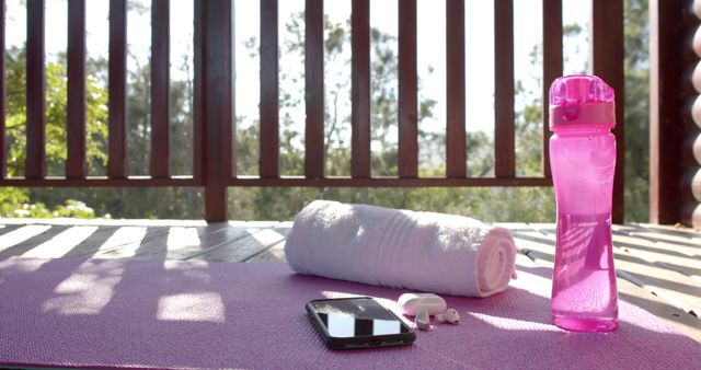 A rolled-up towel, a pink water bottle, a smartphone, and white earbuds sit on a yoga mat on a wooden balcony. Trees and natural light create a serene atmosphere, perfect for at-home or outdoor workout and yoga sessions. Ideal image for promoting fitness routines, exercise equipment, lifestyle blogs, and wellness articles.