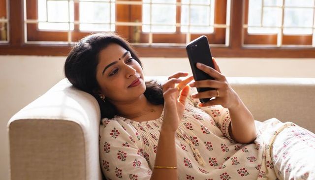 Relaxing woman scrolling smartphone on sunlit sofa wearing floral kurta and bangle