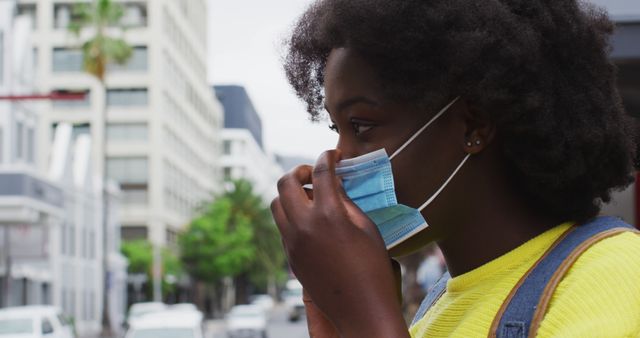 Young woman wearing a yellow top adjusting her face mask on a city street. Tall buildings and green trees line the road, representing urban living during the COVID-19 pandemic. Useful for content related to health precautions, COVID-19 awareness, urban life, and personal safety.