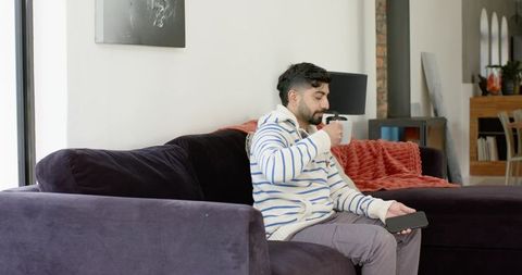 Indian man relaxing on purple sofa drinking coffee while checking smartphone in living room