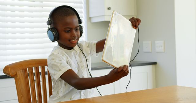 Young boy sits at wooden table at home using a tablet while listening to music with over-ear headphones. He is smiling and seems engaged, suggesting use in contexts such as online learning, technology in education, children enjoying digital devices, or safe online activities for kids.