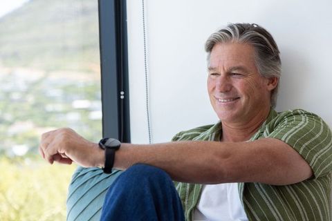 Mature man relaxing on window seat checking wristwatch smiling in casual home lifestyle