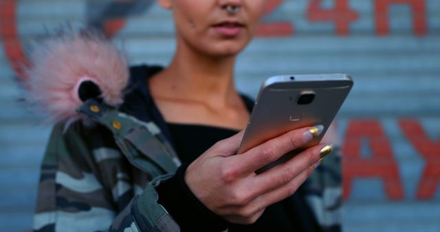 Young woman holding and using a smartphone in an urban setting, wearing a camouflage jacket with faux fur hood detail. The focus is on her hands and smartphone, making it perfect for showcasing mobile technology, urban fashion, or digital communication themes. Ideal for use in advertising, blogs, social media posts, or technology-related articles.