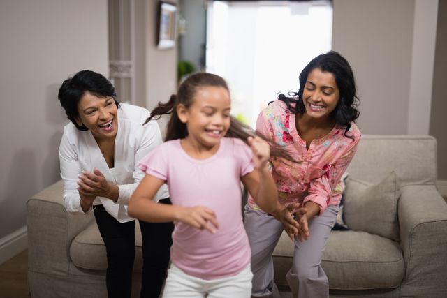 Three generations of a family enjoying a fun moment dancing together in a cozy living room. The child is in the foreground, with the mother and grandmother clapping and smiling in the background. This image can be used for promoting family values, home life, and joyful moments shared with loved ones.