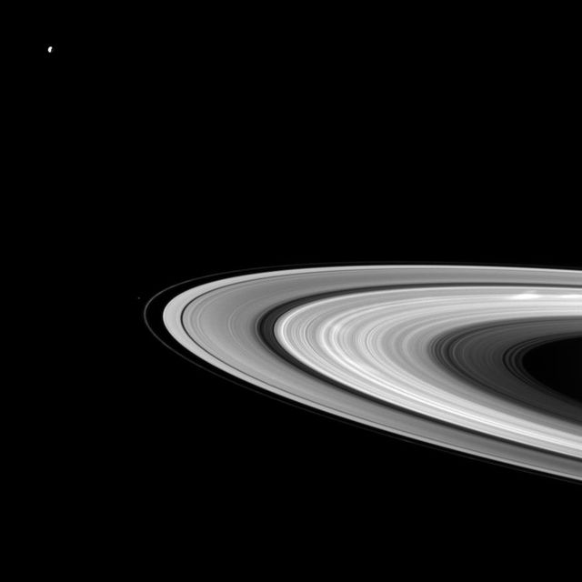 Image shows Saturn's bright rings captured by the Cassini spacecraft, highlighting the planets B ring with visible bright spokes. In the top left corner, Saturns moon Dione stands out, while Pandora appears as a small speck beyond the thin F ring. Ideal for educational content, astronomy enthusiasts, space exploration features, and scientific publications.