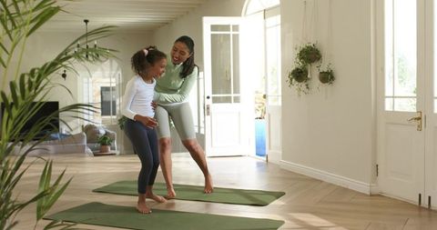 Mother teaching daughter balance on yoga mats in sunlit home living room