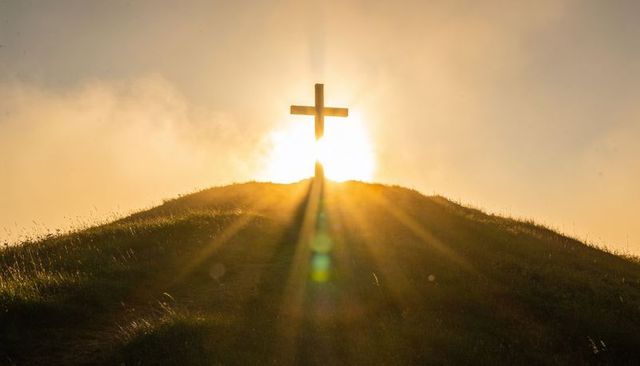 Sunlit cross on hilltop at sunrise with lens flare and warm golden rays