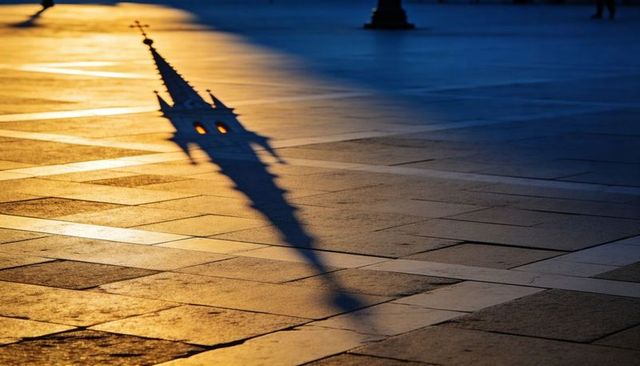 Church spire shadow stretching across tiled plaza at dusk with illuminated lantern windows