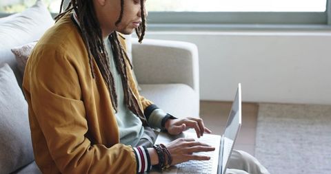 Mid-adult african american man with dreadlocks working remotely on laptop from modern sofa