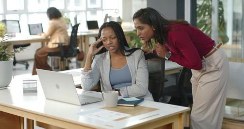 Diverse female coworkers collaborating at laptop in modern open-plan office, teamwork