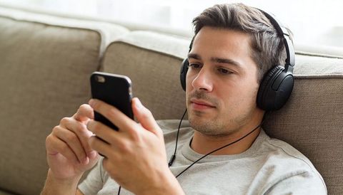 Young man relaxing on couch listening to music with wired headphones and using smartphone