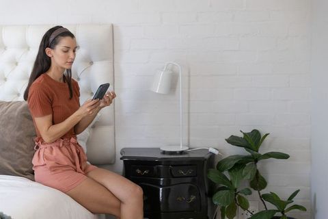 Mid-adult woman sitting on bed using smartphone in minimalist cozy bedroom with plant
