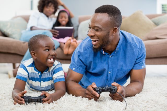 Father and son lying on a rug in the living room, joyfully playing video games together. In the background, mother and daughter are sitting on the couch, engaging with a tablet. This image is perfect for illustrating family bonding, leisure activities, and the use of technology in a home setting. Ideal for use in parenting blogs, family-oriented advertisements, and articles about family time and modern parenting.