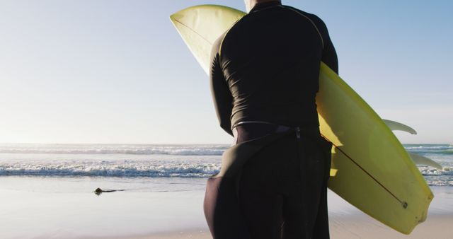 Man holding yellow surfboard stands on sandy beach facing the ocean. Dressed in a wetsuit, he appears to be getting ready for a surfing session. Great for use in articles or advertisements about surfing, water sports, outdoor activities, or beach vacations.