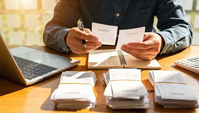Small business owner reconciling receipts at wooden desk with laptop notebook calculator