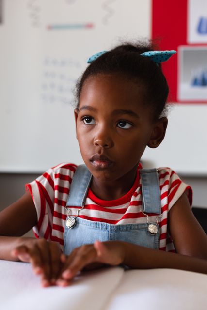 Young African American schoolgirl concentrating while reading a braille book at her desk in a classroom. Ideal for use in educational materials, special education resources, diversity and inclusion campaigns, and articles about disability awareness and inclusive learning environments.