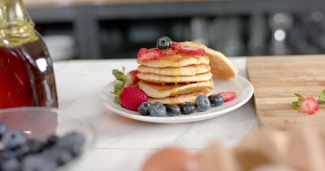 Plate of pancakes garnished with blueberries and strawberries, drizzled with syrup, arranged on marble countertop. Ideal for use in food blogs, breakfast menus, nutrition articles, and restaurant promotions.