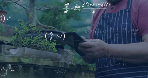 Senior gardener checking smartphone while tending bonsai on stone slabs in striped apron