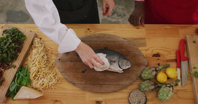Hands of caucasian female cook in black apron preparing fish on cutting board in kitchen. Lifestyle, food, cooking and senior lifestyle, unaltered.