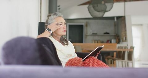 Senior woman sitting on sofa holding tablet rubbing eye with cozy rust blanket in home