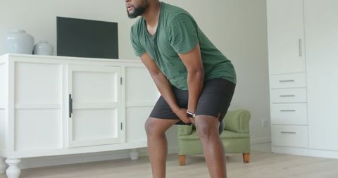 African man bending forward hands on knees wearing smartwatch in minimalist living room