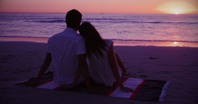 Couple sitting on blanket, watching sunset over ocean, creating a serene and romantic atmosphere. Great for themes around love, relationships, vacations, and relaxation.
