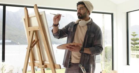 Indian male artist painting on canvas in sunlit waterside studio with beanie headphones
