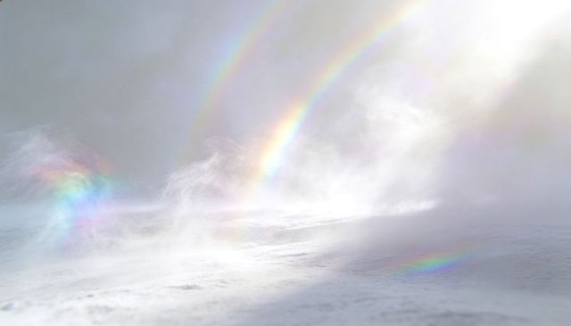 Blowing snow and mist creating vivid double rainbow arcs over icy plateau with prismatic light
