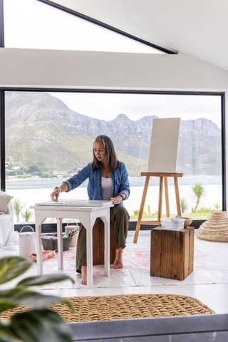 Middle-aged woman painting in bright lakeside studio with mountain view and empty easel