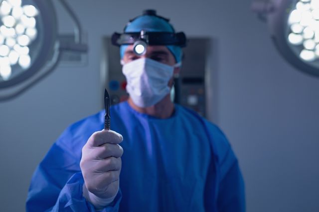 Surgeon in blue surgical gown and mask holding a scalpel in a well-lit operating room. Ideal for use in medical articles, healthcare websites, surgical procedure guides, and hospital promotional materials.