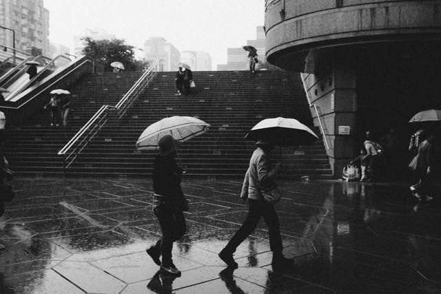 This image shows people walking with umbrellas on a rainy day in an urban setting. The scene captures the essence of daily city life in inclement weather. Useful for themes related to weather, urban life, and transportation in adverse conditions. The black and white tones add a timeless and dramatic effect, making it suitable for artistic and editorial purposes.