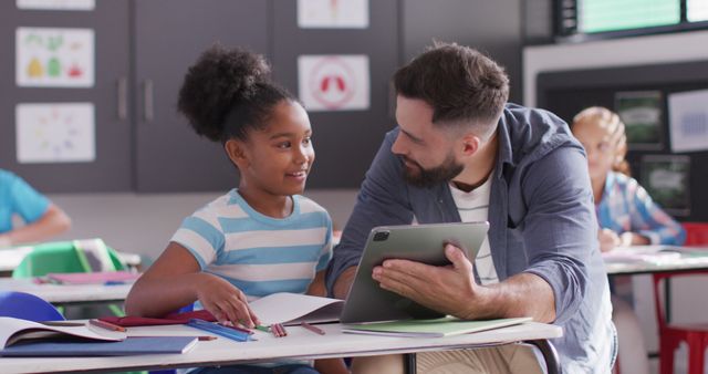 Male teacher using digital tablet with African American student in classroom. Engaged in conversation and collaborative learning. Ideal for use in educational content, articles on technology in education, diverse learning environments, or mentoring programs.