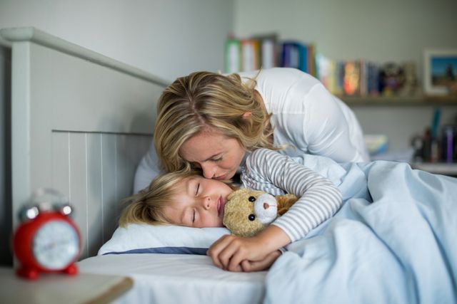 Mother kissing her daughter goodnight while she sleeps in a cozy bedroom. The daughter is holding a teddy bear, creating a warm and loving atmosphere. Ideal for use in parenting blogs, family-oriented advertisements, bedtime routine articles, and content promoting family bonds and affection.