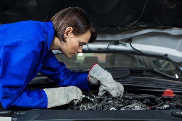 Female mechanic wearing blue jumpsuit and gloves, inspecting a car engine in a repair garage. Suitable for illustrating concepts of women in automotive industry, gender diversity in skilled trades, car repairs, and professional technicians at work.