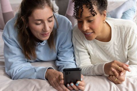Two female friends using smartphone on bed cozy pastel bedroom knitwear lifestyle